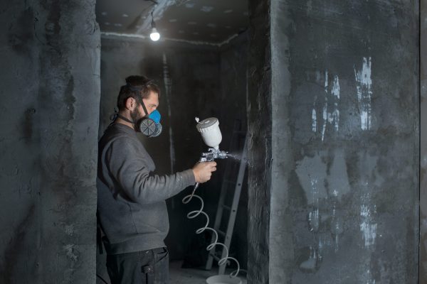 a male plasterer in a construction respirator paints a wall from a spray gun before plastering and putty.
